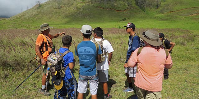 Bowhunter instructor speaking two a group including youth and adults standing in a grassy field.