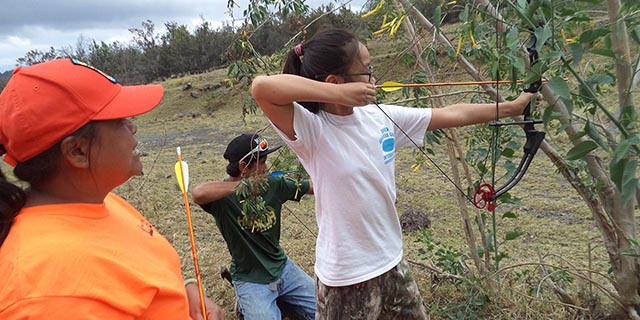 Bowhunter instructor helping a teenage girl and adult male who each is holding bow and arrow.